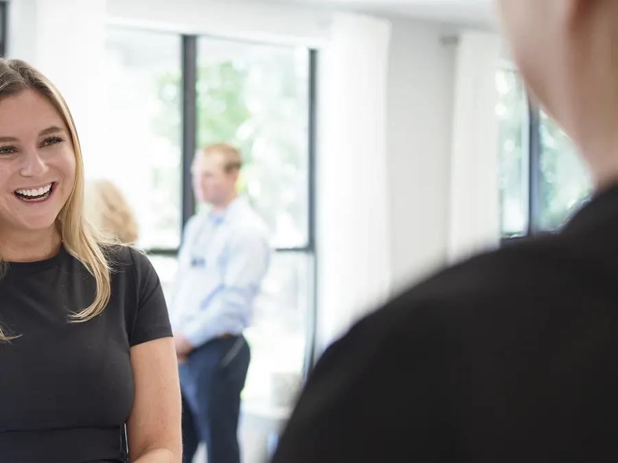 A smiling team member in the practice, with Dr. Perry talking to a patient by the window behind her.